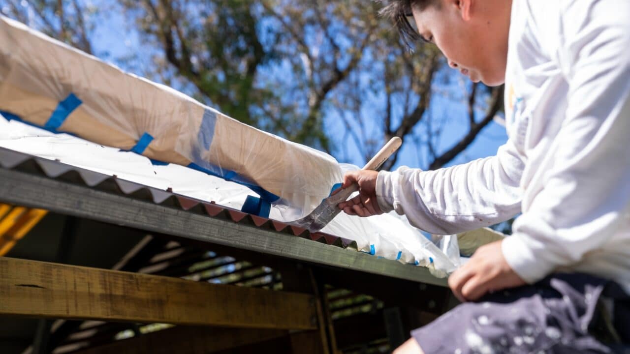 A person is painting corrugated metal roofing while perched on a ladder