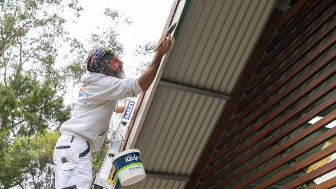 Person wearing white clothing and a colorful headband is standing on a ladder, painting the fascia