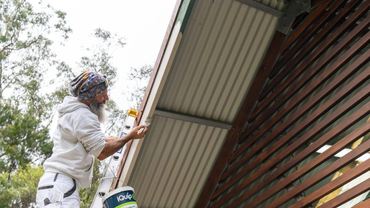 A person on a ladder is painting the eaves of a house while holding a paintbrush