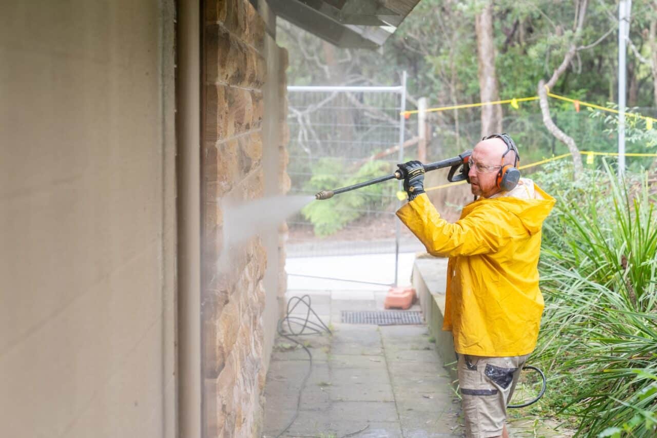 man in a yellow raincoat pressure cleaning a stone wall outdoors