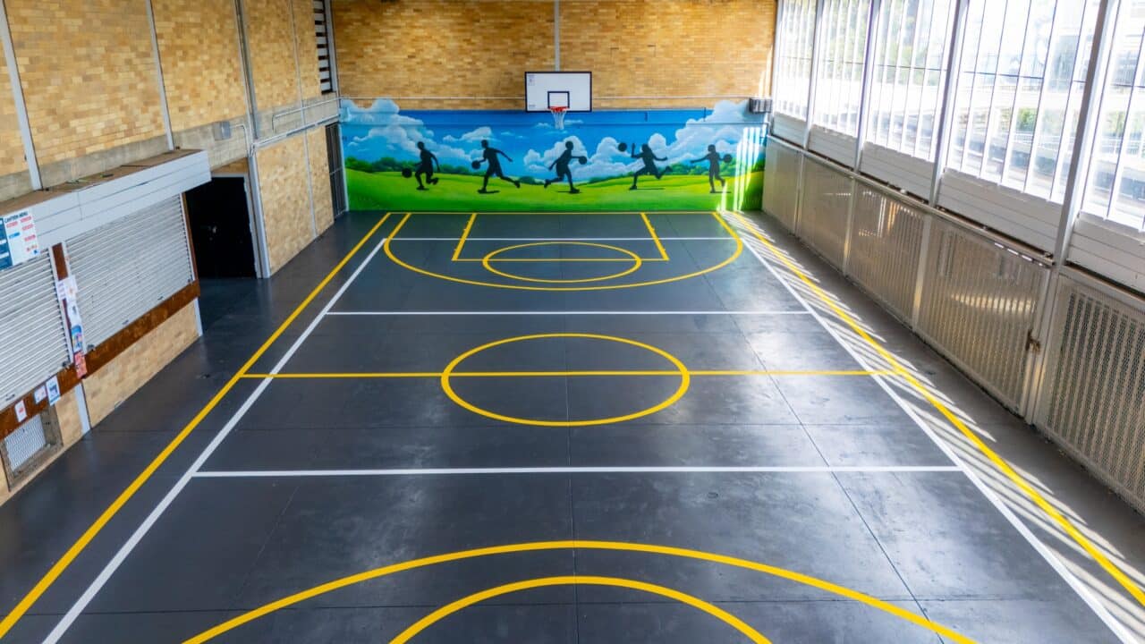the indoor gymnasium at springwood high school features a basketball court with precise line markings in yellow. a mural of people running on grass under a blue sky adorns the far wall, while overhead lights and large windows flood the space with light.