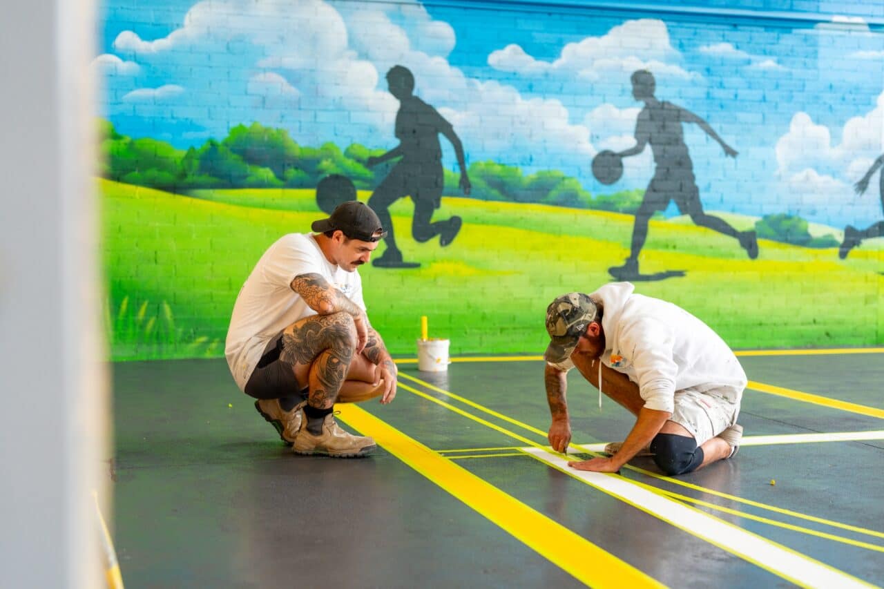 at springwood high school, two individuals in casual attire carefully apply yellow tape for line marking on the gym floor. behind them, a vibrant mural of athletic silhouettes set against a scenic backdrop captures the spirit of the school.