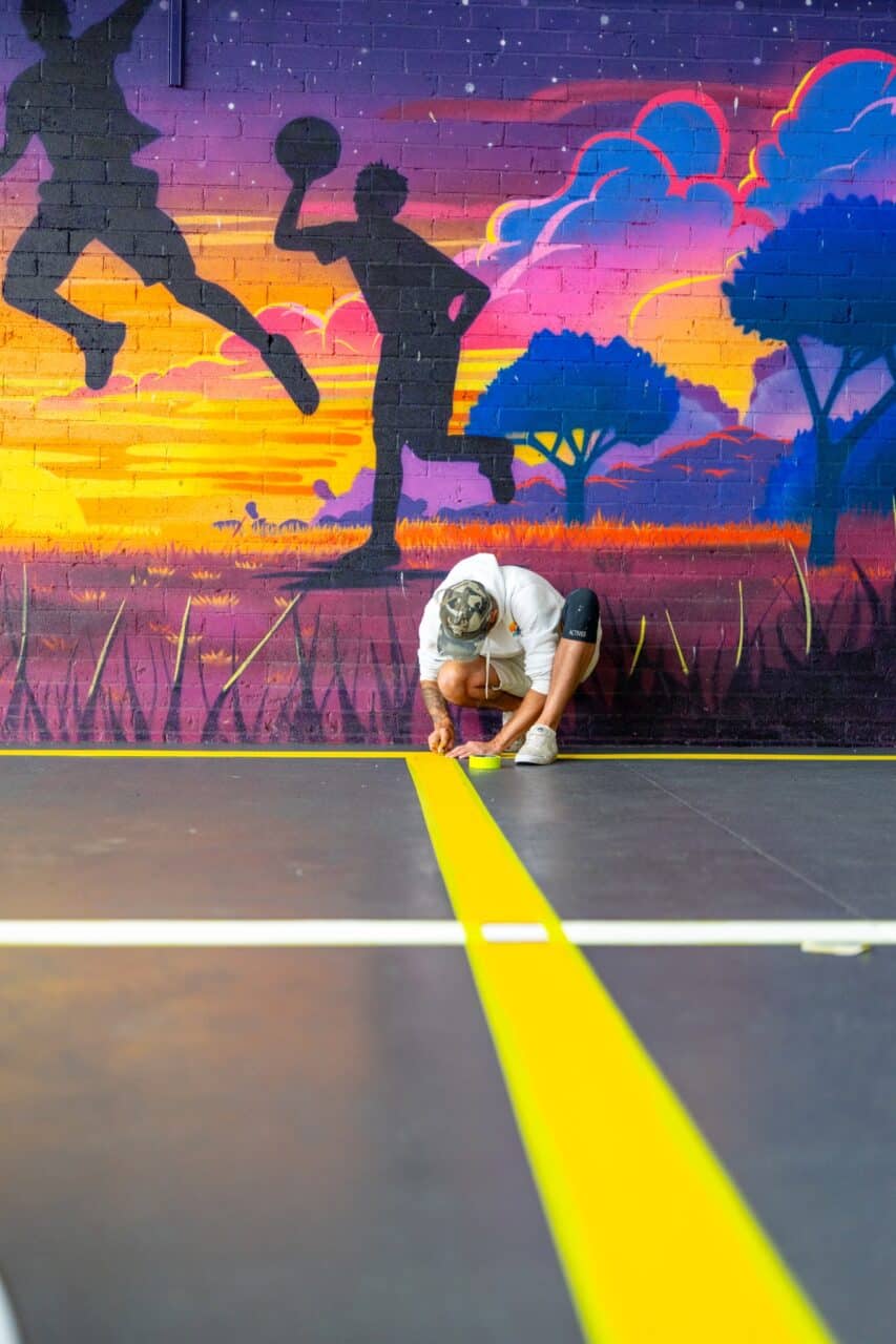 a person in a white shirt and hat is kneeling on the gym floor at springwood high school, carefully applying yellow line marking tape. the background showcases a vibrant mural with silhouettes of athletes against a stunning sunset scene.
