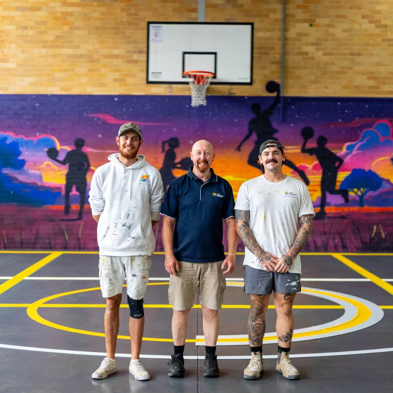 three men standing on the indoor basketball court at springwood high school, with a colorful mural in the background, showcasing silhouettes of people playing basketball at sunset. line marking on the court adds a polished touch to this vibrant setting.