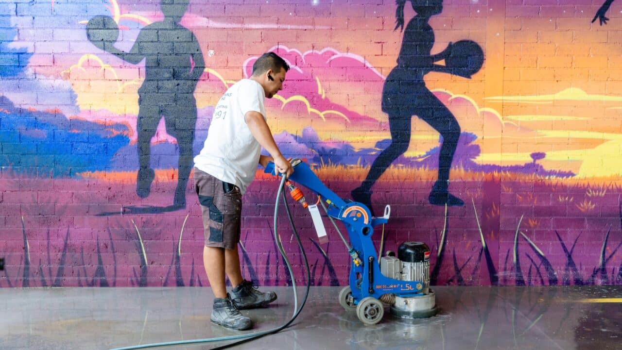 a person operates a floor polishing machine in front of a colorful mural at springwood high school, depicting silhouettes of people playing sports.