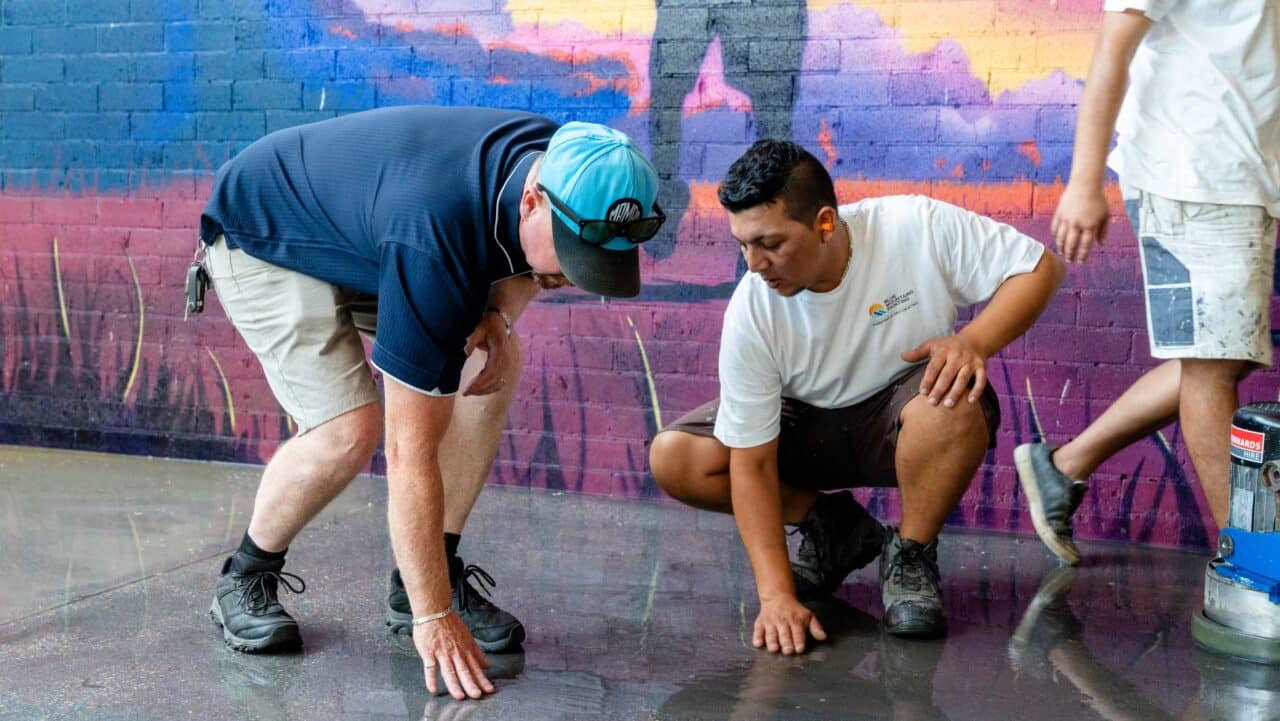 two men kneel on the ground, touching a polished floor at springwood high school. a colorful mural is visible on the brick wall behind them, adding vibrancy to their meticulous line marking efforts.