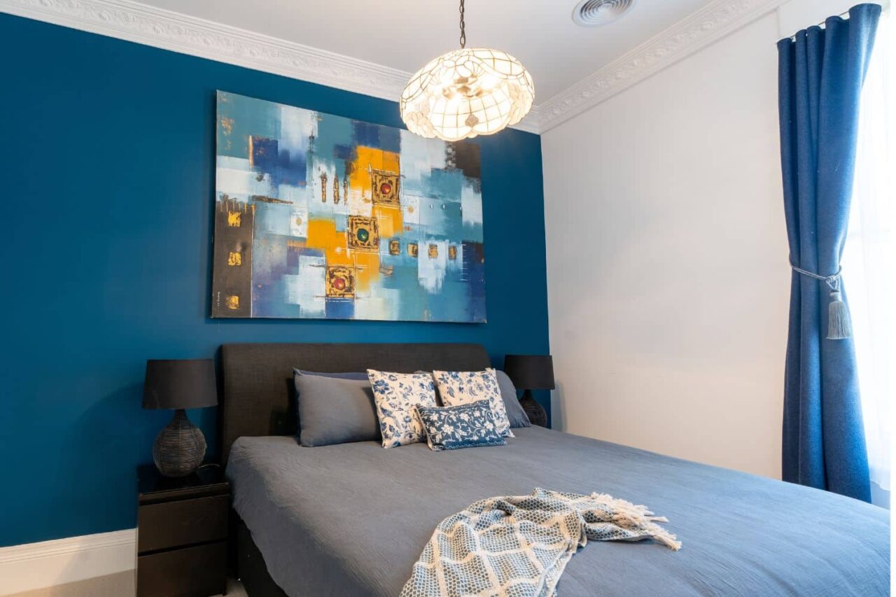 a modern bedroom with a blue accent wall—showcasing the latest interior painting trends—abstract artwork above the bed, two black lamps on nightstands, and blue bedding with patterned pillows and a throw blanket.