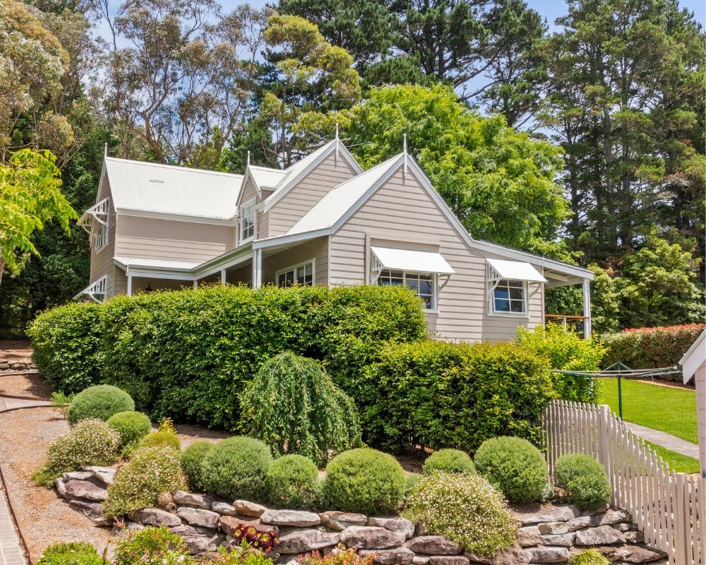 a two story beige house with white trim and a metal roof, surrounded by green bushes, trees, and a landscaped garden with stone edging—a perfect example of home painting excellence by blue mountains painting.