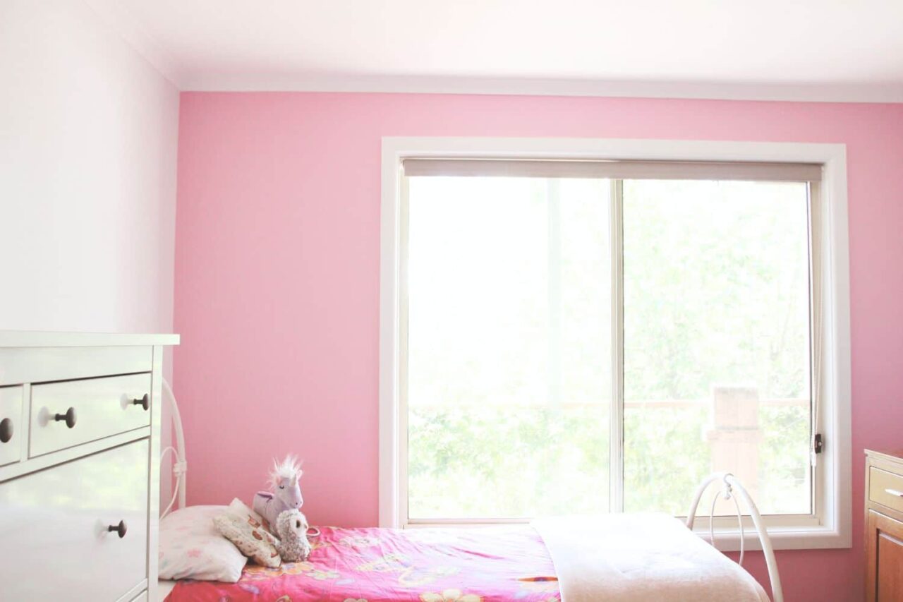 a pink and white bedroom with a bed, stuffed animals, a dresser, and a large window letting in natural light showcases timeless interior paint inspired by the latest interior painting trends.