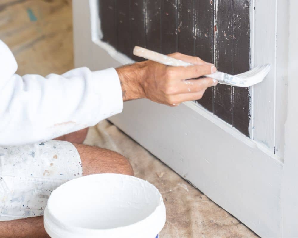 a person paints the edges of a black panel with white paint using a brush; a white paint container sits in the foreground—showcasing home painting with the best paint.