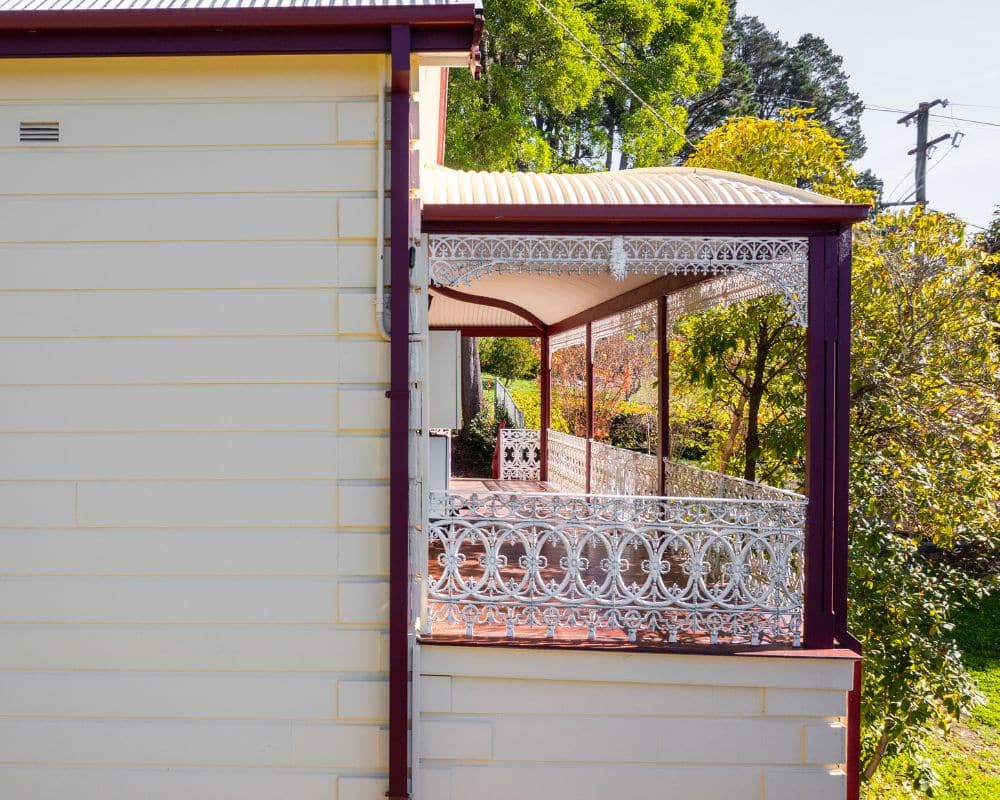 a yellow house with a corrugated metal roof and a decorative white lacework veranda, surrounded by green trees and foliage, evokes the charm of a blue mountains painting.