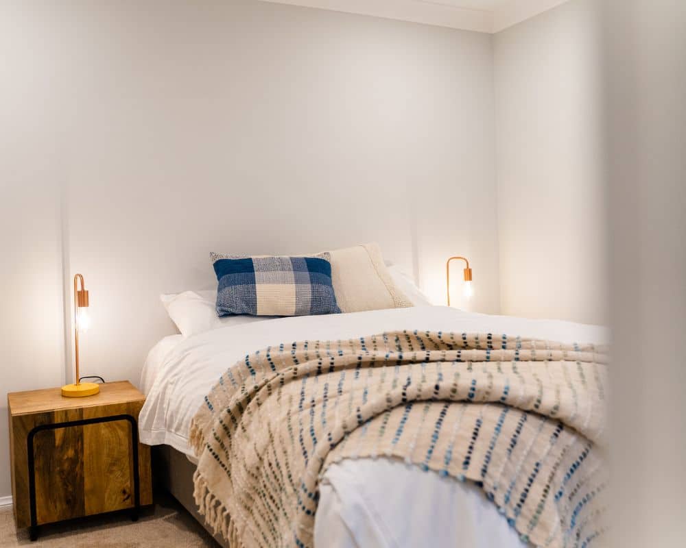 a neatly made bed with white linens, two pillows, and a blue checkered cushion sits beneath a blue mountains painting, flanked by wooden bedside tables and modern lamps in a softly lit bedroom.