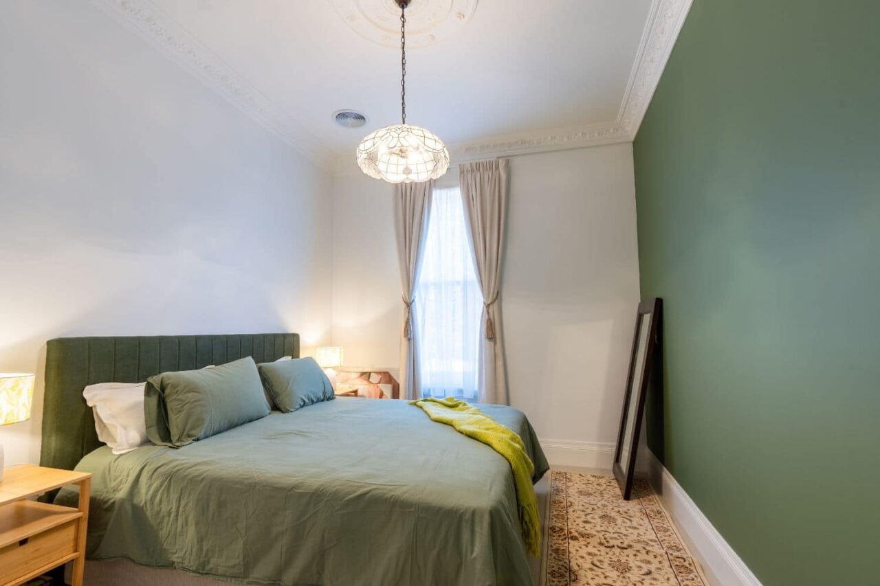a bedroom with a green accent wall showcases timeless trends in interior painting, featuring green bedding, wooden nightstands, a window with curtains, and a floor mirror resting against the wall.