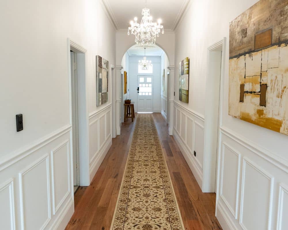 a long hallway with wooden floors, a patterned carpet runner, white paneled walls, modern art including a blue mountains painting on both sides, and two chandeliers hanging from the ceiling.