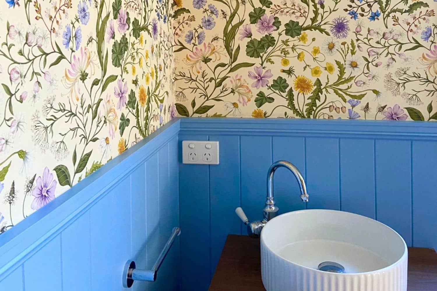 a bathroom with blue wainscoting—a nod to timeless vs temporary painting trends—floral wallpaper, a white round sink, a chrome faucet, and a power outlet above the sink.